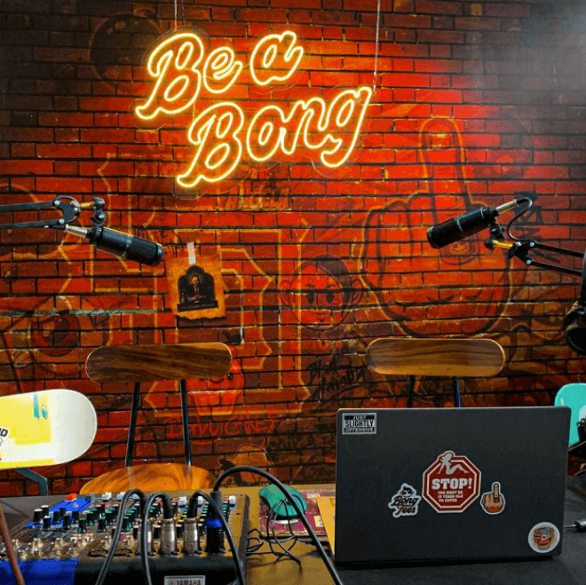 podcast console, microphone and laptop in front of a brick wall with a neon sign